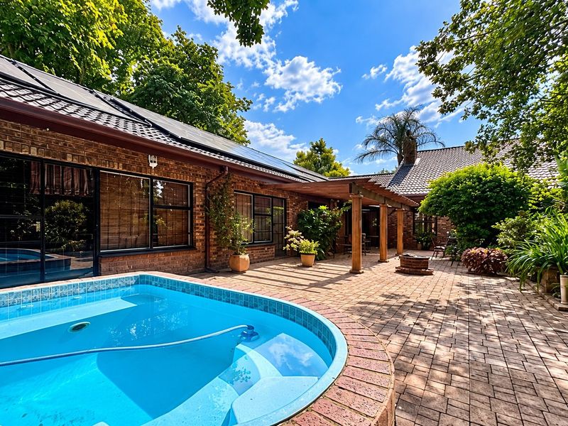A well-maintained outdoor entertainment area featuring a swimming pool, brick-paved patio, and a timber pergola set against a charming brick farmhouse exterior.