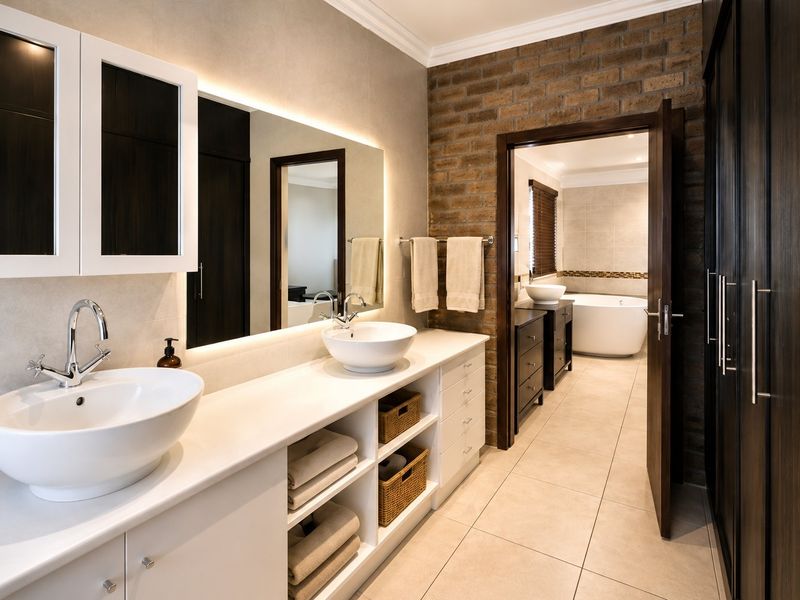 Modern bathroom featuring a double vanity with vessel sinks, backlit mirror, exposed brick accent wall, and a separate area with a freestanding bathtub.