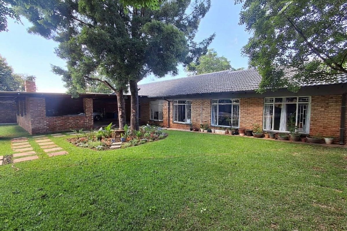 A spacious rear garden featuring a well-maintained lawn, mature trees, a brick-built covered patio area, and a classic face-brick house exterior.