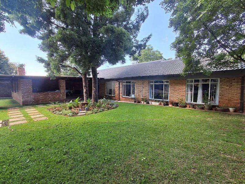 A spacious rear garden featuring a well-maintained lawn, mature trees, a brick-built covered patio area, and a classic face-brick house exterior.