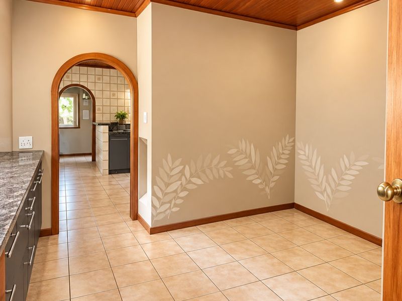 A clean, empty scullery area featuring tiled floors, built-in cabinetry with stone countertops, and a warm timber-paneled ceiling, leading through an archway to the kitchen.