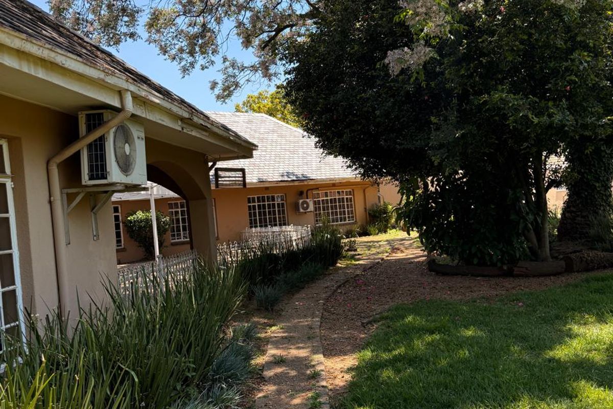 A well-maintained rear garden area featuring a paved walkway, mature trees, and exterior wall-mounted air conditioning units.