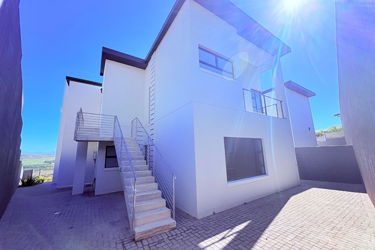 Rear view of a modern multi-level house featuring an external staircase, paved courtyard area, and upper-level balcony with metal railings.