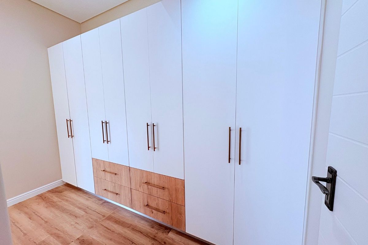 A bright bedroom featuring modern, floor-to-ceiling built-in cupboards with a clean white and wood-grain finish, complemented by durable laminate flooring.