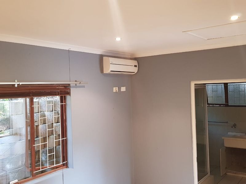 A clean, empty bedroom featuring grey walls, recessed lighting, an air conditioning unit, and a window with security bars, with an adjacent bathroom visible.