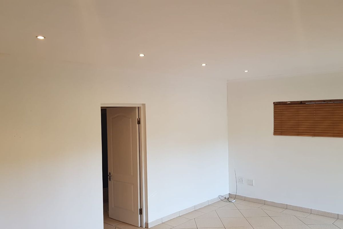 A bright, empty living area featuring neutral tiled flooring, recessed ceiling downlights, and wooden venetian blinds.