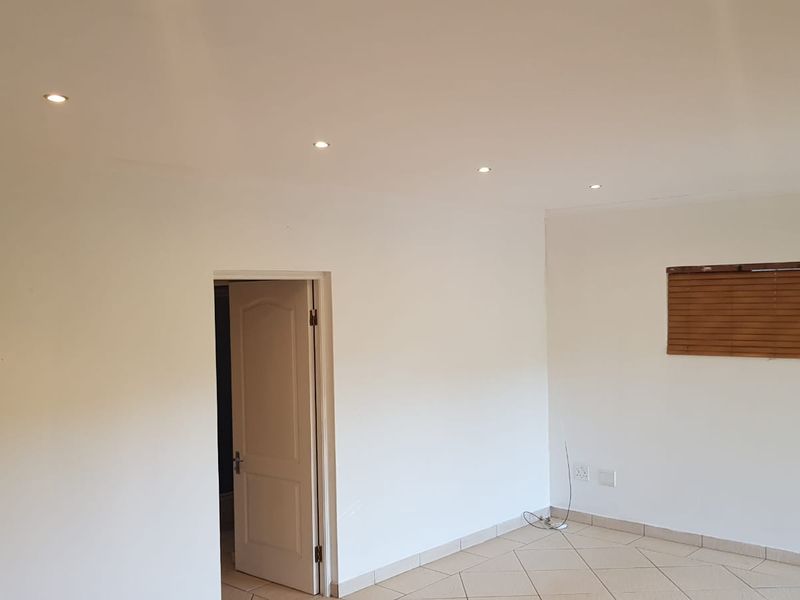A bright, empty living area featuring neutral tiled flooring, recessed ceiling downlights, and wooden venetian blinds.