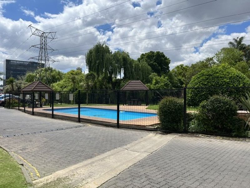 Communal swimming pool area featuring paved surrounds, perimeter fencing, and shaded gazebos within a landscaped garden setting.