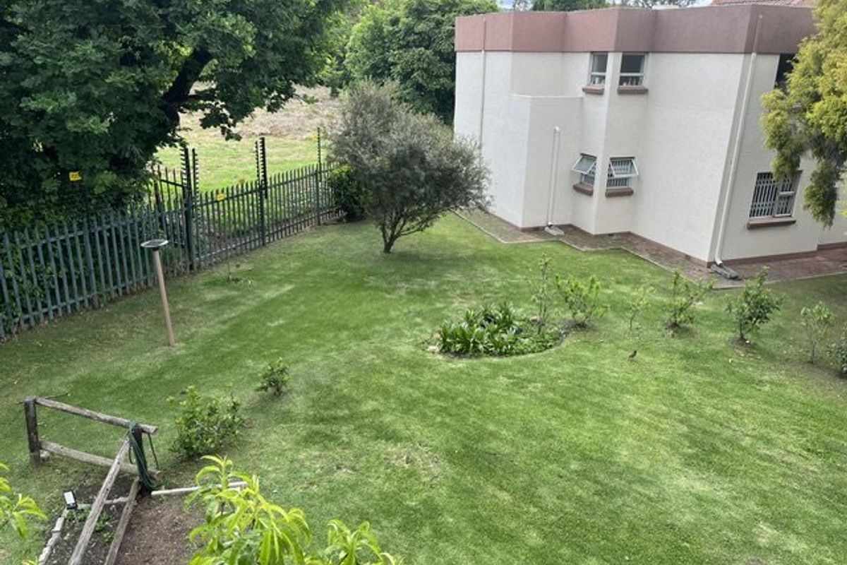 A view of the rear garden area featuring a lawn, established trees, and security fencing surrounding the property perimeter.
