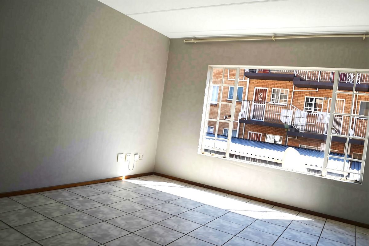 A bright, empty living room featuring tiled flooring and a large window with a curtain rail, offering a view of the neighboring apartment complex.