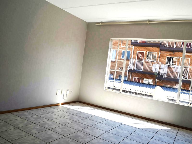 A bright, empty living room featuring tiled flooring and a large window with a curtain rail, offering a view of the neighboring apartment complex.