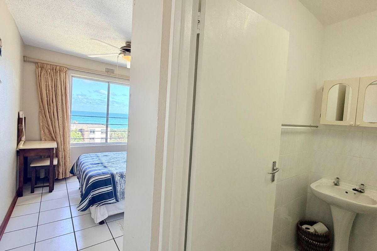 A bedroom featuring tiled floors, a ceiling fan, and an adjoining bathroom area with a pedestal basin and medicine cabinet, offering ocean views.