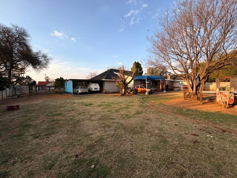 A spacious rear garden area featuring a carport, established trees, and a view of the main house structure with a tiled roof.