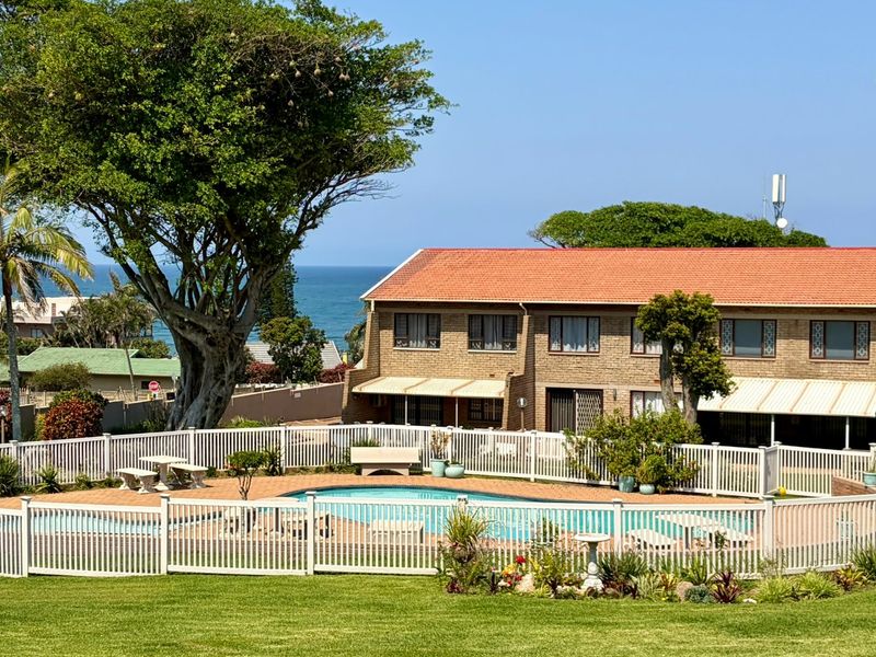 A well-maintained communal pool area featuring paved surrounds, perimeter fencing, and established landscaping, set against a townhouse complex with ocean views.