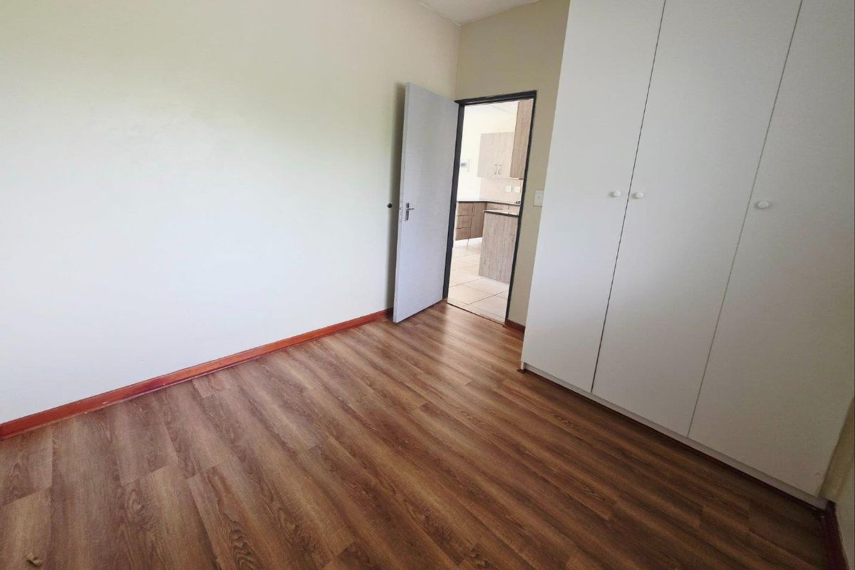 A clean, empty bedroom featuring modern laminate flooring and built-in white cupboards, with a doorway leading to the kitchen area.