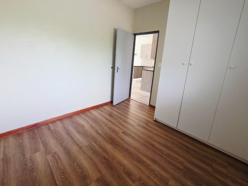 A clean, empty bedroom featuring modern laminate flooring and built-in white cupboards, with a doorway leading to the kitchen area.