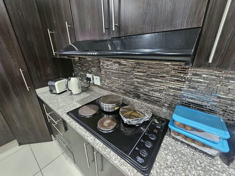 A functional kitchen area featuring dark wood cabinetry, granite countertops, a mosaic tile backsplash, and a built-in electric hob with an extractor fan.
