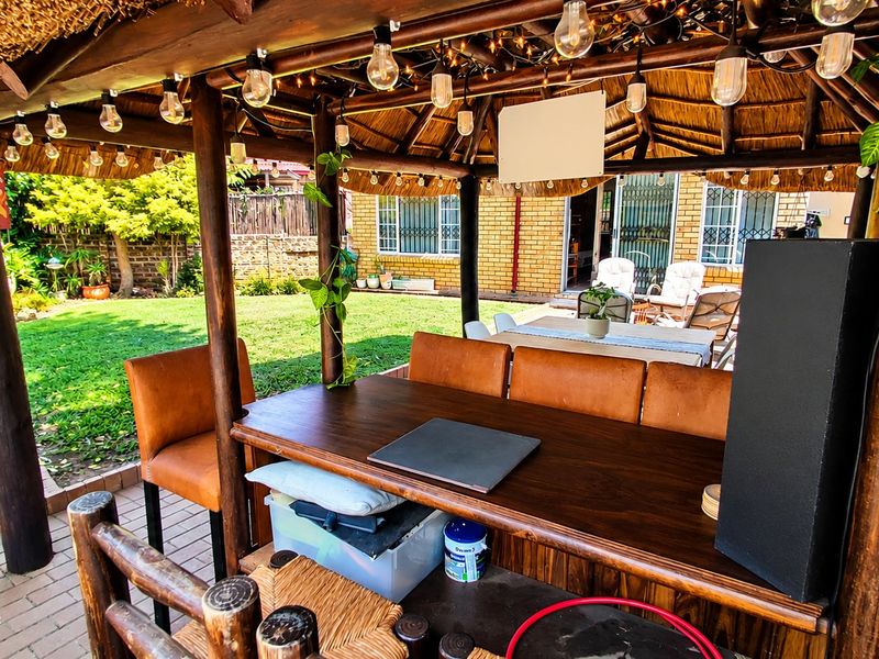 A covered outdoor patio area featuring a thatched roof, timber support posts, and a built-in bar counter, overlooking a landscaped garden.