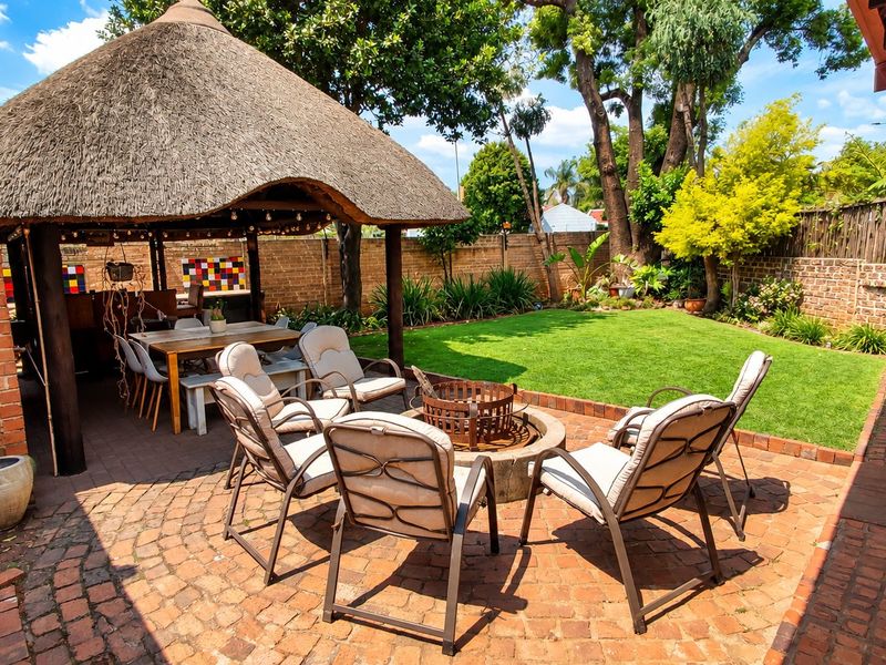 A spacious outdoor entertainment area featuring a traditional thatched lapa, brick-paved patio, and a built-in fire pit set within a private garden.