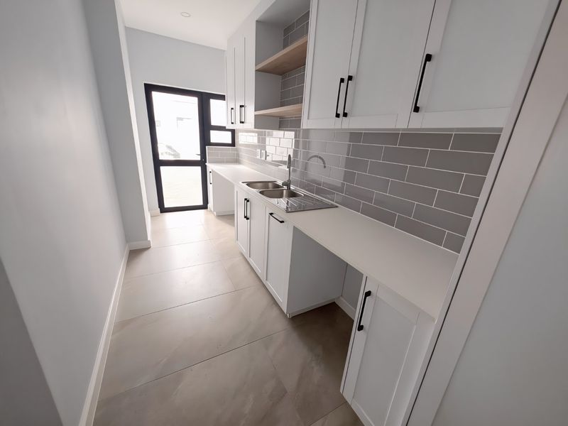 A modern, clean scullery featuring white shaker cabinetry, grey subway tile backsplash, stone countertops, and a stainless steel double sink with direct exterior access.