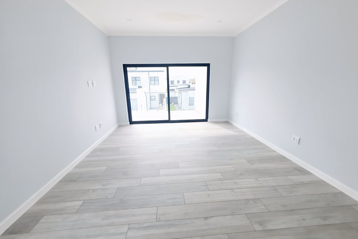 A bright, empty living room featuring modern light-toned laminate flooring, recessed ceiling lighting, and a large sliding glass door providing natural light.