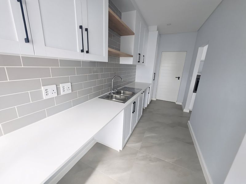 A modern, clean scullery featuring white cabinetry, stone countertops, a double bowl sink, and grey subway tile backsplash with neutral tiled flooring.