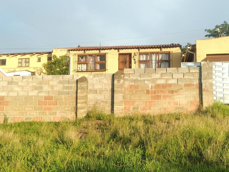 Front view of a single-story house featuring a concrete block boundary wall, metal gate, and tiled roof, set behind an overgrown grass verge.