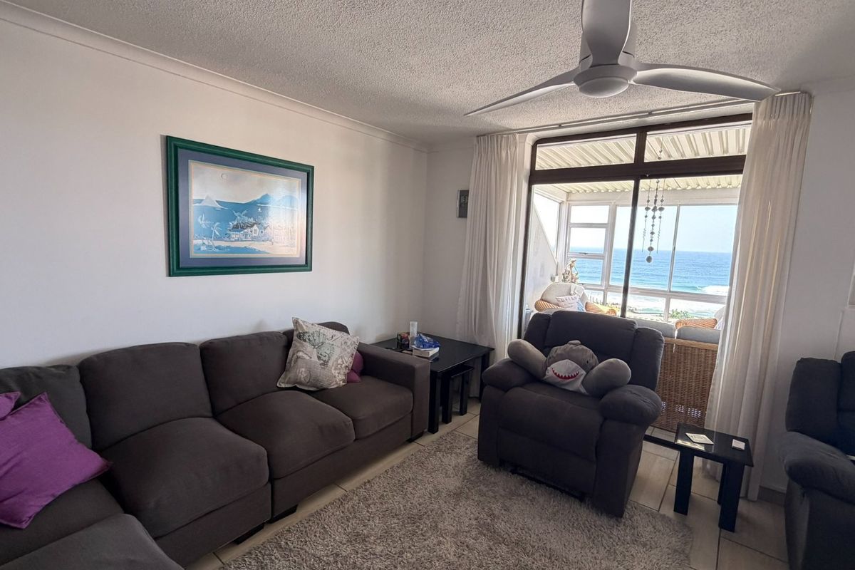 A comfortable living room featuring tiled flooring, a ceiling fan, and large sliding glass doors that provide direct access to a balcony with ocean views.