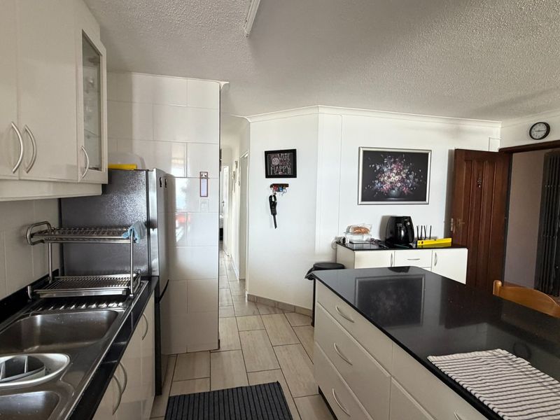 A functional kitchen featuring white built-in cabinetry, dark granite countertops, a double basin sink, and tiled flooring, leading into a hallway.