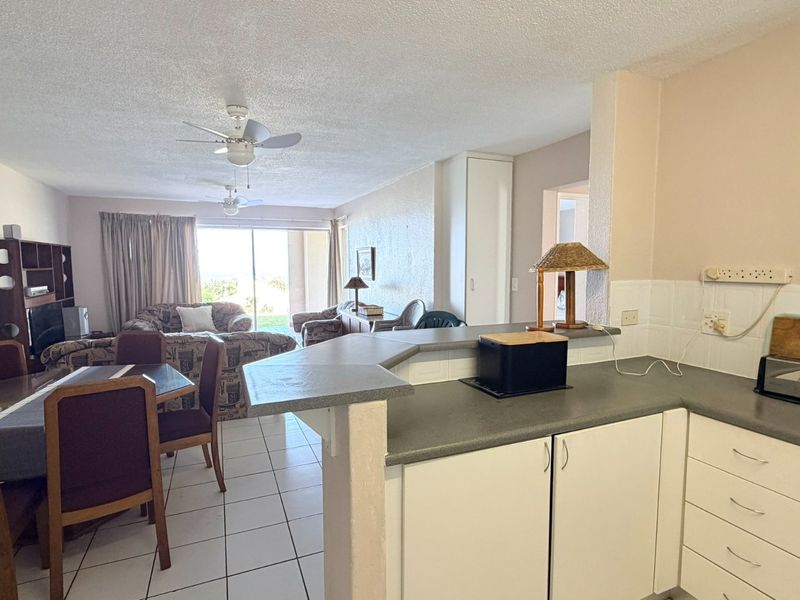 Open-plan living and dining area featuring a functional kitchen with breakfast bar, tiled flooring, and direct access to a patio through sliding glass doors.