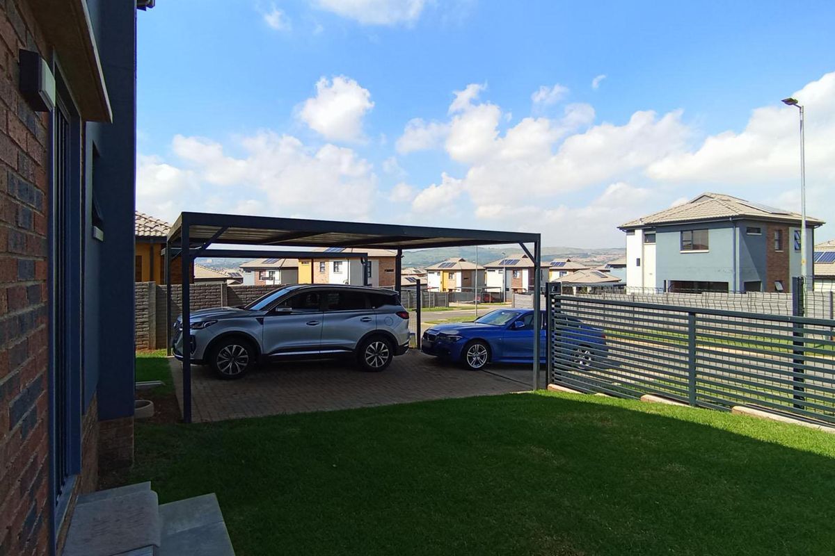A well-maintained rear exterior area featuring a paved driveway with a modern carport, enclosed by metal fencing and a neat lawn.