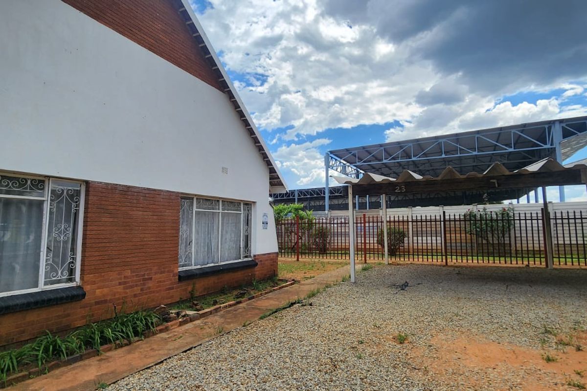 Rear exterior view of the property featuring a brick facade, security-barred windows, and a covered carport area with gravel driveway.