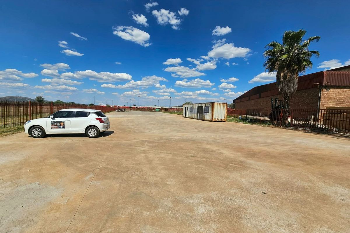 A large, open vacant land plot featuring concrete paving, perimeter steel fencing, and a shipping container structure.