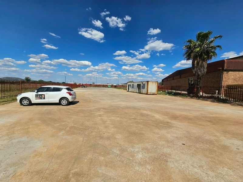 A large, open vacant land plot featuring concrete paving, perimeter steel fencing, and a shipping container structure.