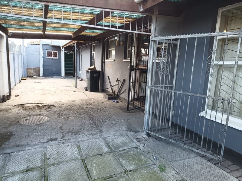 Rear exterior area featuring a covered patio, security gate, and paved driveway with visible wear to the concrete surfaces and exterior wall finishes.