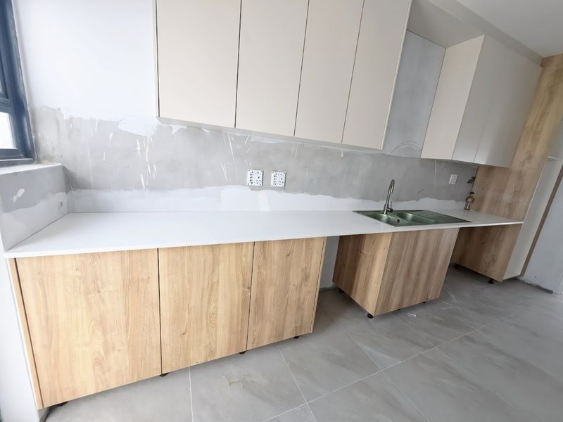 A modern kitchen in progress featuring light wood-look cabinetry, white stone countertops, and a stainless steel sink, awaiting final wall finishes and splashback installation.