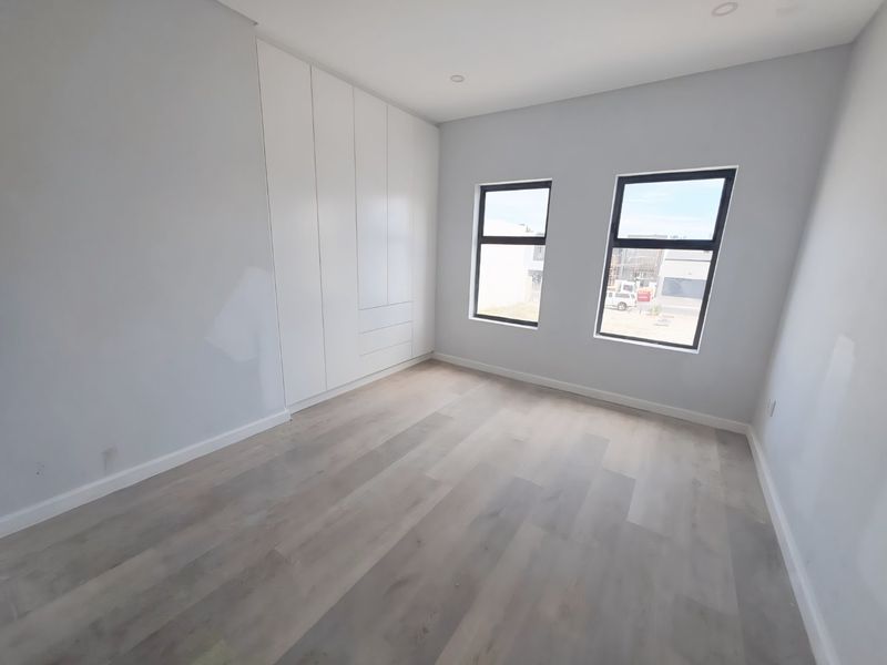 A bright, modern, and empty bedroom featuring light-toned laminate flooring, built-in white cupboards, and large windows with aluminium frames.