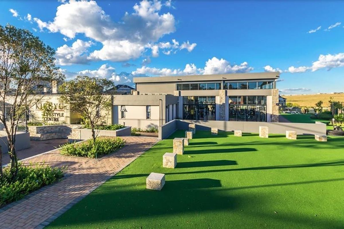 A well-maintained exterior area featuring a paved walkway, artificial lawn, and stone seating, leading to a modern building with a large glass facade.