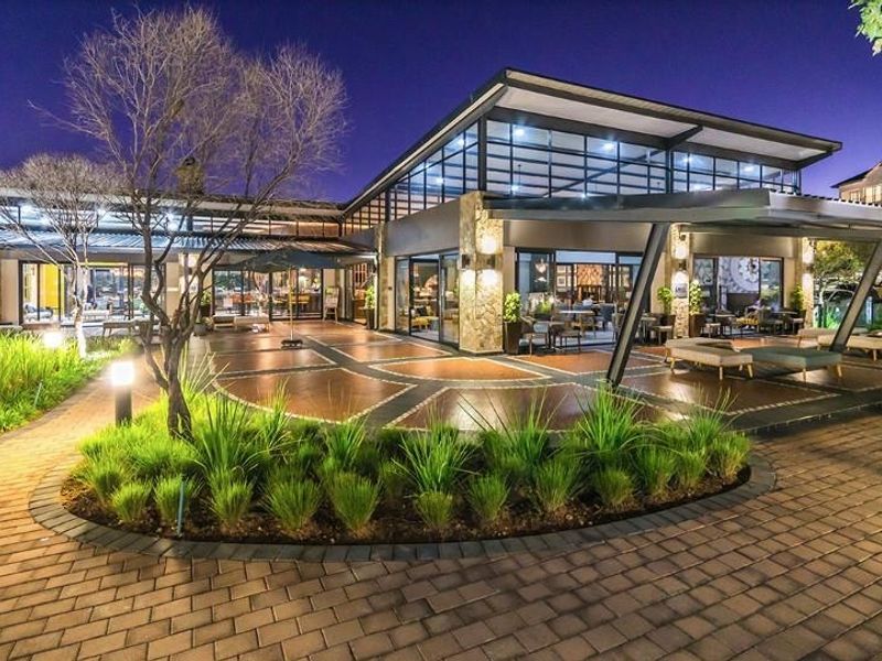 A modern clubhouse exterior featuring a spacious paved courtyard, stone-clad pillars, and extensive glass facades, illuminated for evening use.