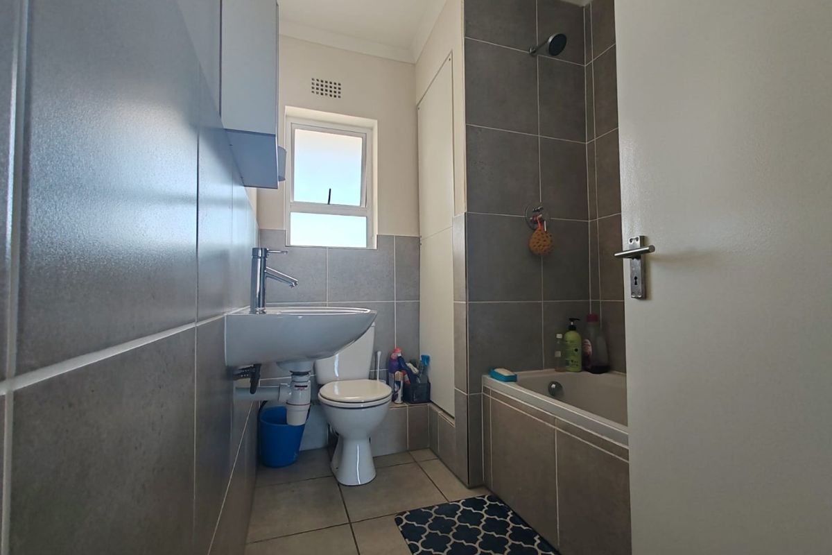 A compact, modern bathroom featuring grey wall tiles, a wall-mounted basin, a toilet, and a bathtub with an integrated shower head.