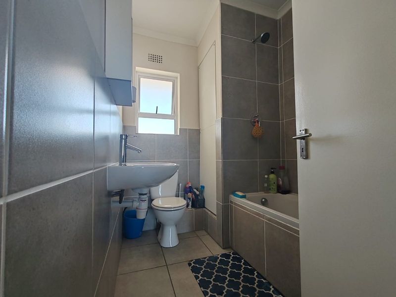 A compact, modern bathroom featuring grey wall tiles, a wall-mounted basin, a toilet, and a bathtub with an integrated shower head.
