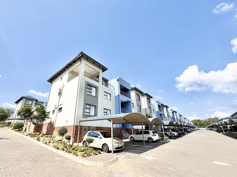 Exterior view of a modern multi-story residential complex featuring paved parking areas, covered carports, and balconies with aluminium window frames.