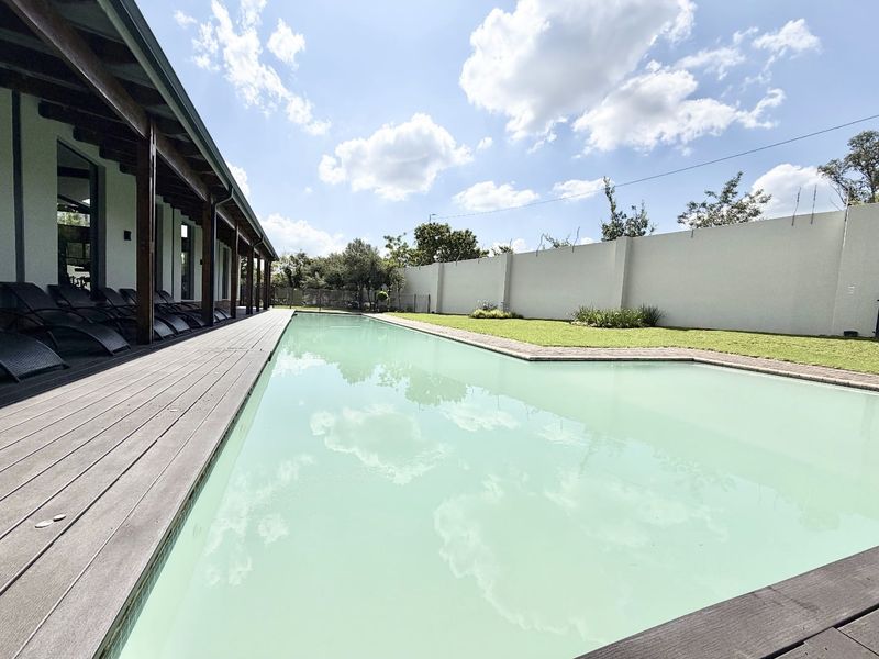 A spacious swimming pool area featuring extensive timber decking, a covered patio, and a well-maintained lawn enclosed by a high perimeter wall.