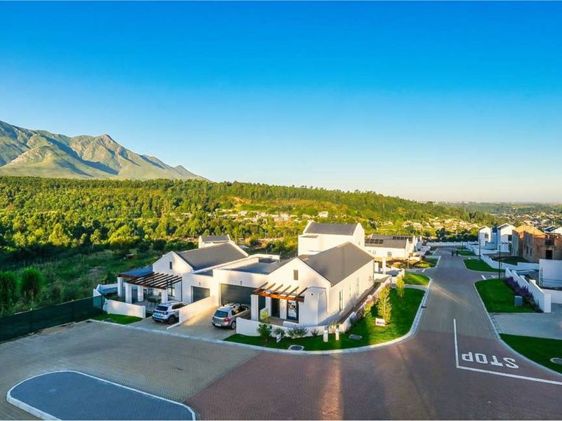 Aerial view of a modern residential estate featuring contemporary homes with pitched roofs, timber pergolas, and paved driveways set against a scenic mountain backdrop.