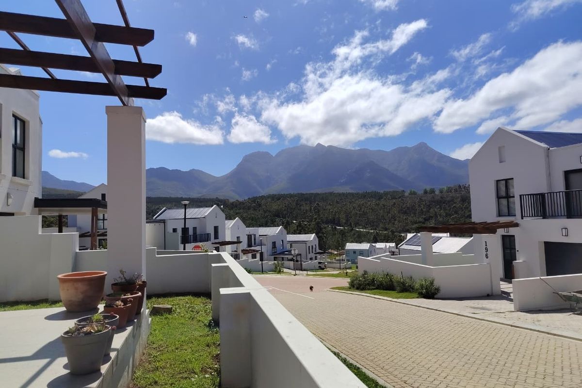 A modern residential exterior view featuring a paved driveway, white building facades, and a timber pergola, set against a scenic mountain backdrop.