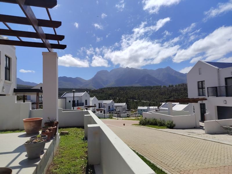 A modern residential exterior view featuring a paved driveway, white building facades, and a timber pergola, set against a scenic mountain backdrop.