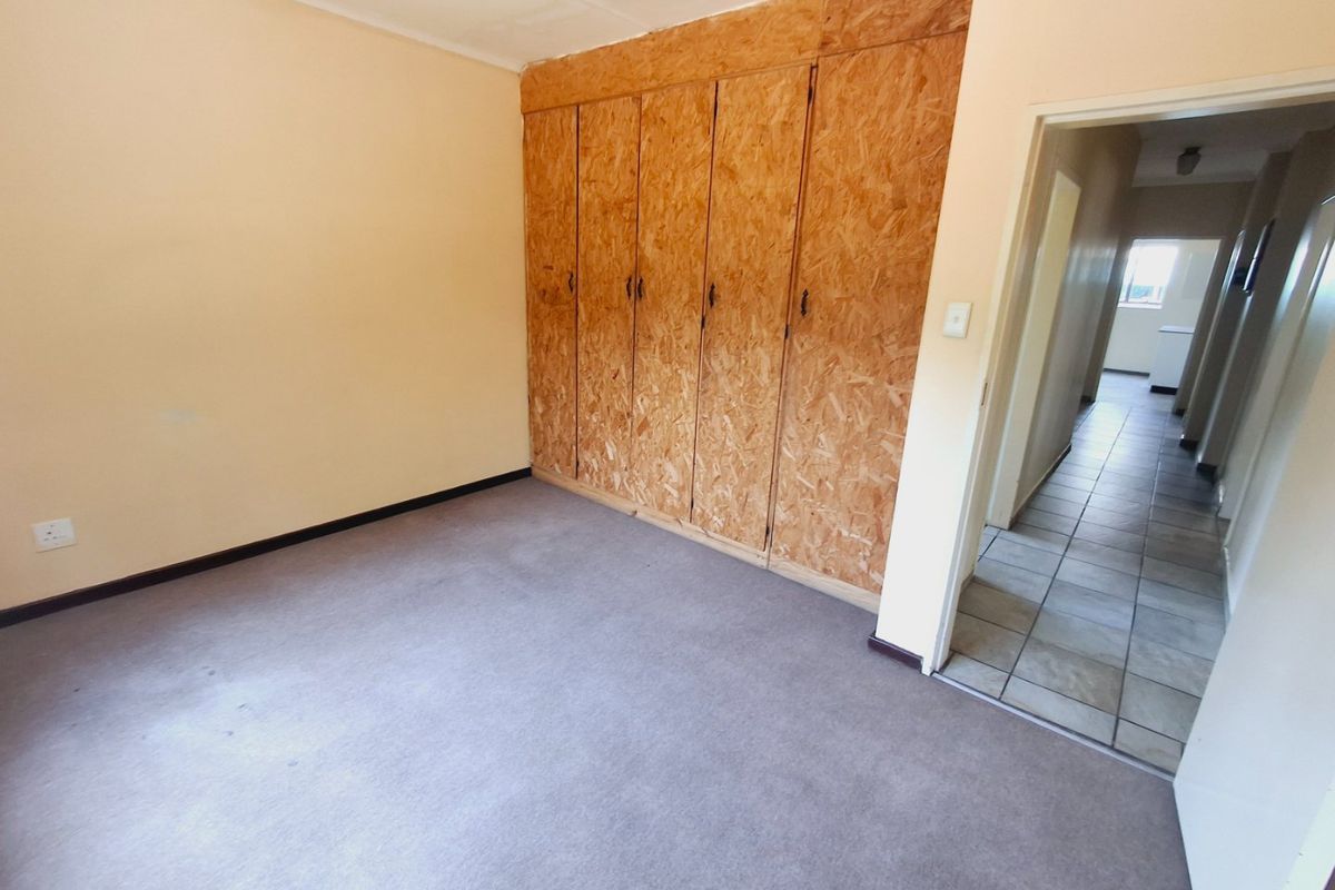 An empty bedroom featuring built-in cupboards with a raw wood finish and carpeted flooring, leading into a tiled hallway.