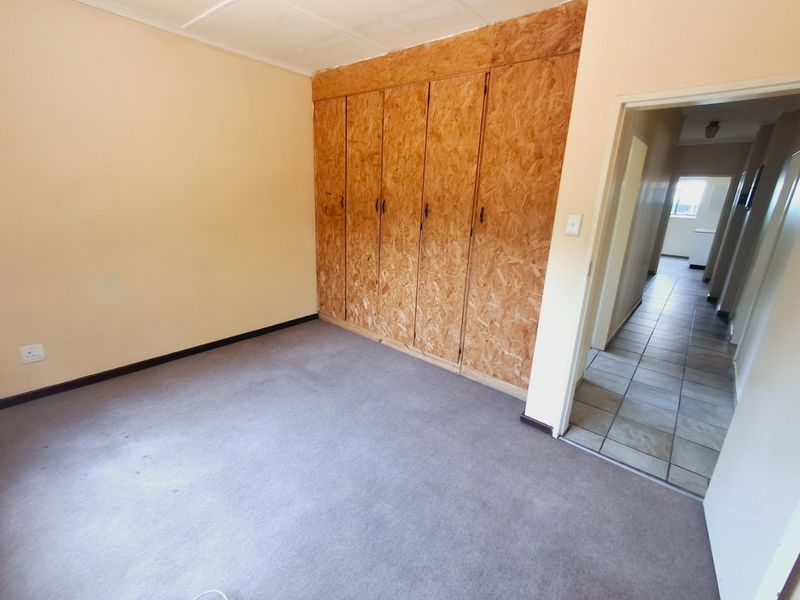 An empty bedroom featuring built-in cupboards with a raw wood finish and carpeted flooring, leading into a tiled hallway.