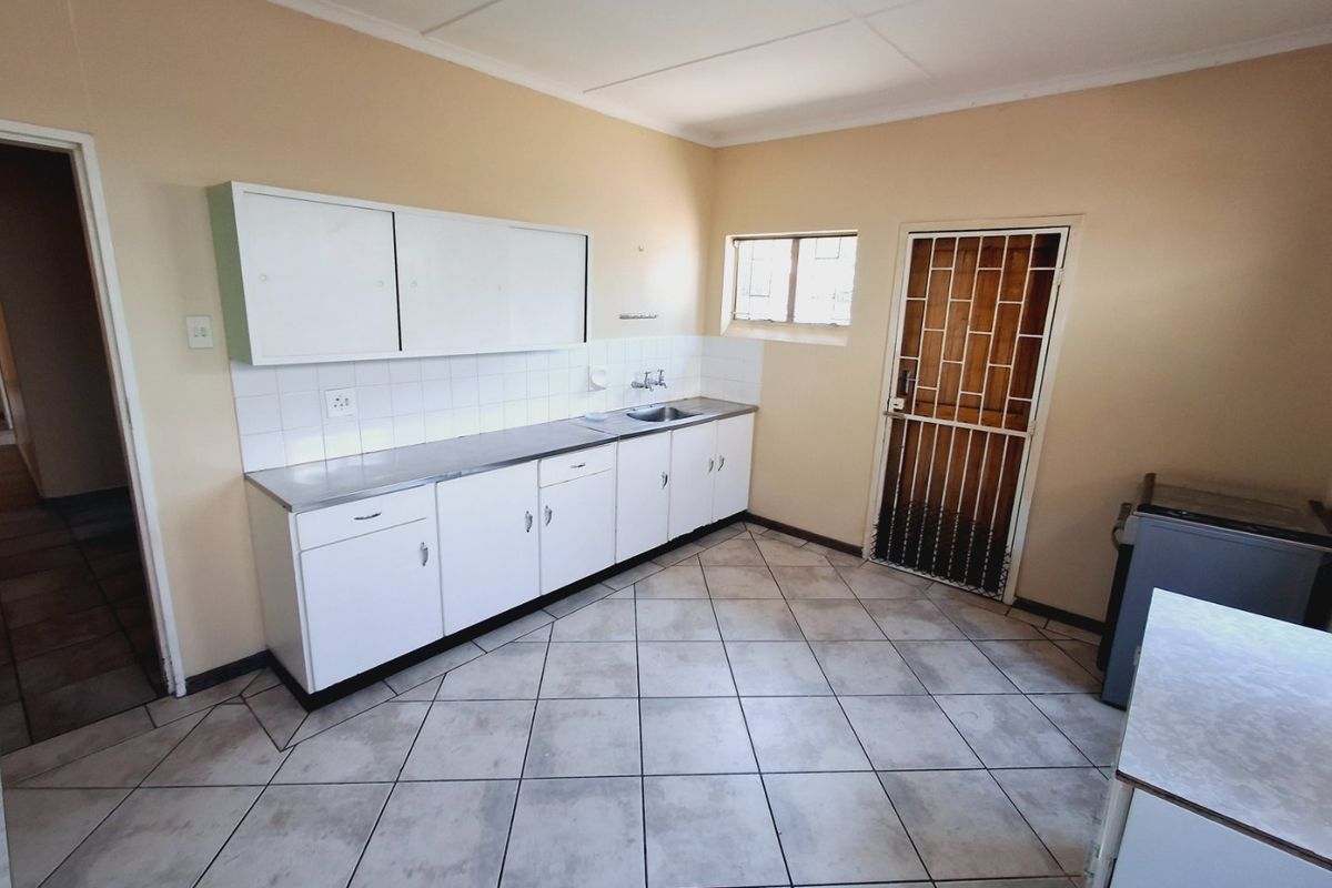 A functional kitchen featuring built-in white cabinetry, a stainless steel sink, tiled splashbacks, and durable floor tiles with direct access to an exterior security door.