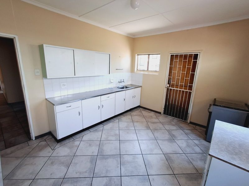 A functional kitchen featuring built-in white cabinetry, a stainless steel sink, tiled splashbacks, and durable floor tiles with direct access to an exterior security door.